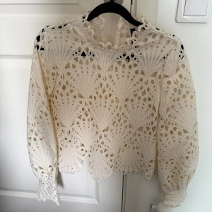 Vit broderad blus från JDY - Superfin vit blus från JDY med broderade detaljer och genombrutet mönster. Blusen har långa ärmar med resår vid ärmslut och dragkedja i ryggen. 