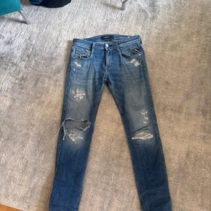 Blå slitna jeans från Replay - Säljer ett par blå skinny jeans från Replay med snygga slitningar och hål på benen. Jeansen har klassisk femficksdesign, låg midja och är tillverkade i mjukt denimtyg. Perfekta för dig som gillar en avslappnad och trendig look.