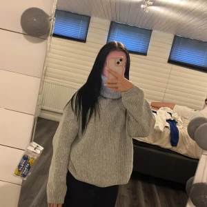 Grå ribbad polotröja i stickat - Mysig grå polotröja i stickat material med ribbad struktur. Tröjan har en oversized passform och långa ärmar, perfekt för kyliga dagar. Hög polokrage som ger extra värme och stil. Enkel att matcha med både jeans och leggings.