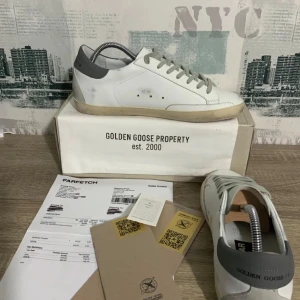 Golden Goose vita sneakers med grå detaljer - Golden Goose sneakers i vitt skinn med grå snörning och grå hälkappa. Klassisk stjärndetalj i mocka på sidan och GGDB/STAR-tryck. Sulan är beige och lätt slitad för en vintage-look. Perfekta för dig som gillar streetstyle och exklusiva märken.