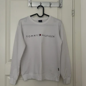 Vit sweatshirt från Tommy Hilfiger - Vit sweatshirt från Tommy Hilfiger med klassisk logga tryckt framtill. Tröjan har rund halsringning, långa ärmar och ribbade muddar. Perfekt för en clean och stilren look. Materialet känns mjukt och skönt mot huden. Pris går att diskuteras.