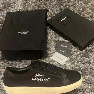 Svarta Saint Laurent sneakers i skinn - Svarta sneakers från Saint Laurent med vit handskriven logga på sidan, guldfärgad logga på plösen och häl, samt vit platt sula. Skorna är i skinn och textil, har snörning och en klassisk rund tå. Snygg minimalistisk design med exklusiv känsla.