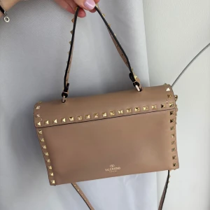Beige Valentino axelväska med nitar - Säljer en beige axelväska från Valentino med ikoniska guldiga nitar runt kanterna och på axelremmen. Väskan är i slätt skinn och har både handtag och lång axelrem. Invändigt finns flera fack och dragkedja. Perfekt för dig som gillar edgy men stilren design.