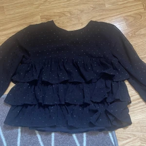 Svart volangblus från Gina Tricot - Supersöt svart blus från Gina Tricot med långa ärmar och flera lager volanger. Blusen har små glittriga prickar över hela tyget och en knapp i nacken. Perfekt för dig som gillar en lite extra look!