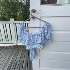 Blommig croppad blus med knyt - Supersöt blommig croppad blus i vitt och blått med puffärmar och knyt i ryggen. Perfekt för en somrig look!💙