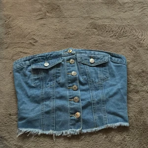 Jeans tubtopp med knappar och fransad kant - Säljer en snygg jeans tubtopp med knappar framtill, två fejkade bröstfickor och fransad nederkant. Baksidan har smock för extra stretch och passform. Perfekt för dig som gillar denim och vill ha en cool, edgy look. Storlek S. Knappt använd, skicket är som nytt 