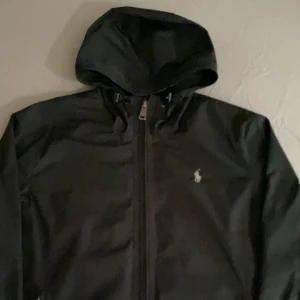 Svart vindjacka från Polo Ralph Lauren - Svart vindjacka från Polo Ralph Lauren med huva och dragkedja framtill. Jackan har en stilren look med diskret logga på bröstet och är tillverkad i ett lätt och smidigt material. Perfekt för blåsiga dagar och enkel att matcha med olika outfits.