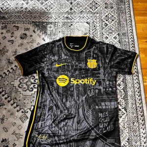 Barcelona x Nike svart fotbollströja - Snygg Barcelona fotbollströja från Nike i svart med gula detaljer och tryck. Tröjan har korta ärmar, gul Nike-logga, Spotify sponsor och klubbmärke i gult. Materialet är lätt och andas, med ett unikt grafiskt mönster över hela tröjan. Perfekt för dig som älskar Barca!