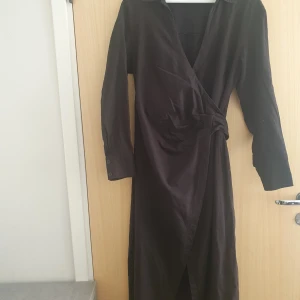 Svart omlottklänning i linne - Svart lång omlottklänning med knyt i midjan och långa ärmar. Klänningen har klassisk krage, knappar vid ärmslut och är tillverkad i svalt linnetyg. Klänningen är helt ny och aldrig använd 