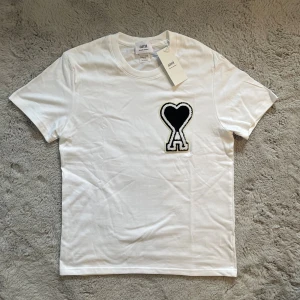Vit t-shirt Ami Paris - 🖤 Klassisk vit T-shirt med den ikoniska svarta Ami de Cœur-loggan på bröstet.🖤 Ny med taggar kvar.  📏 Storlek: M ✅ Skick: Helt ny / oanvänd                                       🚚 Frakt: Samma / nästa dag                                        DMa för frågor!