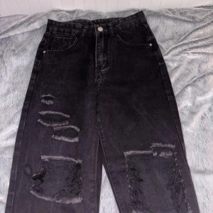 Svarta ripped jeans med raka ben - Säljer ett par svarta jeans med snygga slitningar och hål på båda benen. Modellen har raka ben och klassisk femficksdesign med knapp och dragkedja. Jeansen är tillverkade i kraftigt denim och har en cool, edgy vibe som passar streetstilen.🖤