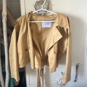 Beige trenchcoat från Bershka S 😍 - Snygg beige trenchcoat från Bershka i kortare modell med klassisk krage och bälte i midjan. Jackan har långa ärmar, knappar framtill och en clean look som passar perfekt till lager-på-lager. Materialet känns mjukt och lätt, perfekt för vår och höst!