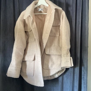 Beige overshirt från NA-KD, stl 34 - Säljer en beige overshirt från NA-KD i storlek 34. Jackan har fyra stora fickor framtill, klassisk krage och knappar vid ärmslut. Materialet är mjukt och borstat, perfekt för lager-på-lager under höst och vår.