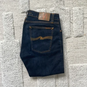 Mörkblå skinny jeans från Nudie Jeans - Säljer ett par mörkblå slim jeans från Nudie Jeans med klassiska orange sömmar och snygg vågig detalj på bakfickan. Jeansen har normal passform i midjan och är tillverkade i denim för extra komfort. Perfekta för dig som gillar en slimmad look.