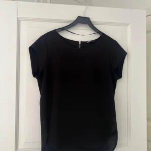 Svart blus med kort ärm - En stilren svart blus med rund halsringning och korta ärmar. Blusen har en enkel och klassisk design med lätt rundad nederkant och dragkedja bak i nacken. Perfekt att matcha med både jeans och kjol.