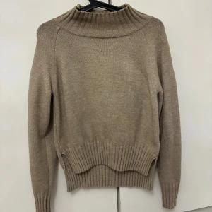Beige stickad polotröja - Beige stickad polotröja med bred ribbad krage och muddar. Tröjan har lång ärm och en relaxed passform, perfekt för lager-på-lager. Enkel och stilren design som funkar till många olika outfits.