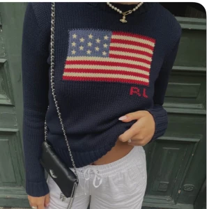 Mörkblå polotröja från Ralph Lauren - Mörkblå stickad polotröja från Ralph Lauren med amerikansk flagga framtill och RL-broderi. Tröjan har lång ärm, ribbade muddar och hög polokrage. Perfekt för dig som vill ha en klassisk men ändå cool look. Tröjan är storlek Xxs-S