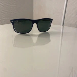 Blå Ray-Ban Wayfarer solglasögon - Snygga Ray-Ban Wayfarer solglasögon i mörkblå plast med gröntonade glas. Klassisk fyrkantig form och diskret Ray-Ban-logga på bågarna. Perfekta för att lyfta din stil under soliga dagar. Fodral medföljer.
