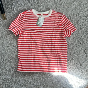 Rödvit randig t-shirt  - Säljer en fräsch t-shirt från H&M med röda och vita horisontella ränder. T-shirten har rund hals med vit kant och korta ärmar. Skön passform och tillverkad i mjuk bomull, perfekt för chill dagar. Passar dig som gillar klassisk streetstil.