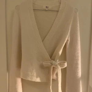 Vit/beige omlottkofta från Gina Tricot - Supermysig vit/beige omlottkofta från Gina Tricot i ullblandning. Koftan har långa ärmar, ribbade kanter och knyts i sidan med ett brett band. Perfekt för lager på lager och chill dagar.