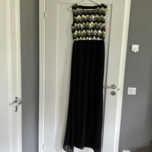 Svart långklänning med paljetter Dry Lake - Snygg långklänning från Dry Lake med svart kjol och överdel täckt av glittriga paljetter i guld, silver och svart i ett zickzackmönster. Ärmlös modell med rund halsringning, perfekt för att sticka ut på festen.