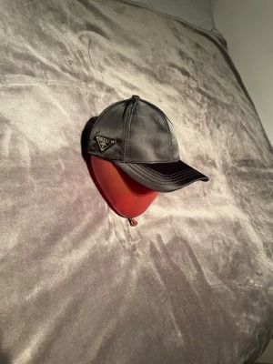 Svart keps från Prada - Snygg svart keps från Prada med glansig satinfinish och klassisk böjd skärm. Kepsen har justerbar rem baktill och en ikonisk Prada-logga på sidan. Perfekt för dig som vill ha en stilren accessoar med lyxig känsla.