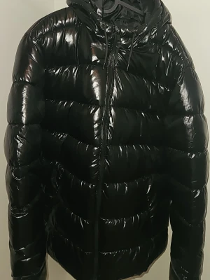 Svart glansig pufferjacka med huva - Säljer en svart pufferjacka med glansig finish och huva. Jackan har dragkedja framtill och breda, vadderade kanaler som ger en riktigt puffig look. Perfekt för kalla dagar när du vill hålla värmen och ändå se cool ut.