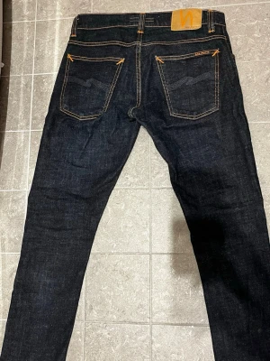 Nudie Jeans W28 L30 - Säljer ett par mörkblåa skinny jeans från Nudie Jeans i storlek W28 L30. Jeansen är tillverkade av 100% ekologisk bomull och har klassiska kontrastsömmar samt fem fickor. Snygg passform med smala ben och stilren look.