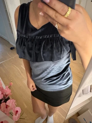 Mörkblå sammetslinne med volang - Säljer ett mörkblått linne i mjuk sammet med bred volangdetalj över bröstet. Toppen är ärmlös men man kan även lägga till armarna eftersom de är volanger och har en lätt glansig finish som ger en lyxig känsla. Perfekt att styla med kjol eller jeans för en trendig look.