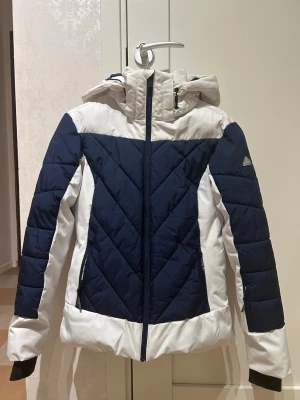 Skidjacka från McKinley - Mörkblå och vit McKinley skidjacka i storlek 36. Jackan har snölås och flera innerfickor samt ficka för liftkort på armen. Jackan har tecken på användning då det finns spår av smink på insidan av kragen som jag tyvärr inte fått bort i tvätten.  Jackan är väldigt varm har använts upp till 20 minus grader.  Armlängd från axeln: 60 cm