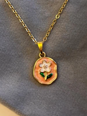 Gulligt Halsband Blomma guld imitation  - Så sött halsband med en berlock av en blomma mot en färgad bakgrund. Berlocken är ca 1 x 1,3 cm.Trädd på kedja i guldimitation, justerbar 40-45 cm. 