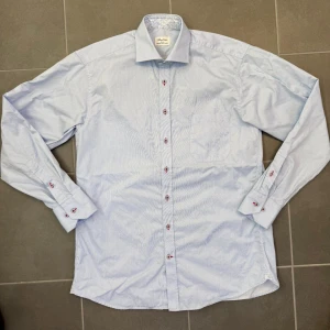 Ljusblå randig skjorta från Stenströms stl 40 - Säljer en klassisk ljusblå skjorta från Stenströms med tunna vita ränder och röda detaljerade knappar. Skjortan har lång ärm, bröstficka och snyggt mönstrat tyg på insidan av kragen. Tillverkad i mjuk bomull för en clean och fräsch look.