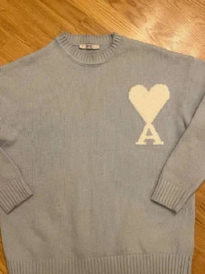 Ljusgrå stickad tröja från Ami helt ny oanvänd. - Ljusgrå stickad tröja från Ami med rund halsringning och ribbade muddar. På bröstet finns ett stort vitt hjärta med bokstaven A under. Perfekt för dig som gillar stilrena och trendiga plagg. STORLEK L-XL