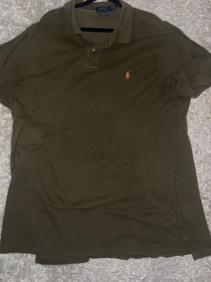 Olivgrön pikétröja från Polo Ralph Lauren - Säljer en olivgrön pikétröja från Polo Ralph Lauren i storlek XXL. Tröjan har klassisk krage, två knappar och den ikoniska broderade loggan i orange på bröstet. Slim fit-modell i mjuk bomull som är skön att ha på sig hela dagen.
