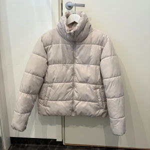Gråbeige pufferjacka med hög krage - Säljer en gråbeige pufferjacka med hög krage och dragkedja framtill. Jackan har två sidofickor och är quiltad för extra värme. Perfekt för kyliga dagar och har en clean, modern look som passar till det mesta.