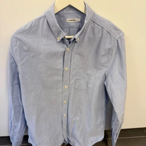 Ljusblå skjorta från J.Lindeberg - Klassisk ljusblå skjorta från J.Lindeberg med button down-krage och bröstficka. Skjortan har långa ärmar, vita knappar och är tillverkad i bomull. Perfekt för en clean och stilren look. Storlek M