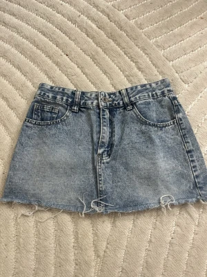 Blå jeanskjol med fransig kant - Säljer en kort jeanskjol i ljusblå tvätt med rå, fransig kant nertill. Kjolen har klassiska fem fickor, bälteshällor och dragkedja framtill. Perfekt för dig som gillar en avslappnad och trendig stil.