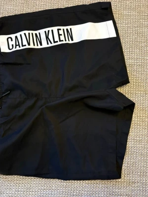 Svarta shorts från Calvin Klein - Svarta shorts från Calvin Klein med vit logga framtill. De har elastisk midja och en bakficka. Materialet känns lätt och syntetiskt, perfekt för varma dagar. Snygg och enkel design som funkar till många stilar.
