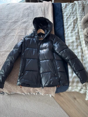 Svart pufferjacka från Save The Duck - Säljer en svart pufferjacka från Save The Duck i storlek M. Jackan är tillverkad i syntetmaterialet Plumtech, vilket gör den både lätt och varm. Den har huva, två dragkedjefickor och hel dragkedja framtill. 100% animal free och vattenavvisande. 