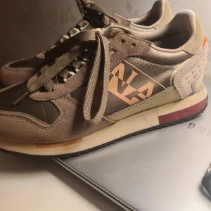 Napapijri sneakers i beige och brunt - Snygga sneakers från Napapijri i beige och bruna toner med stora logodetaljer på sidan. Ovandelen är i mocka och syntet med coola kontraster och snörning. Skorna har en platt sula och rund tå, perfekt för en avslappnad streetstil.