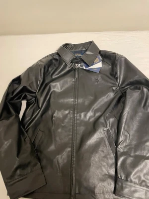 Polo Ralph Lauren jacka - Svart skinnjacka från Polo Ralph Lauren med klassisk krage och dragkedja framtill. Jackan har diskret broderad logga på bröstet och två sidofickor. Insidan har ett rutigt foder. Perfekt för dig som gillar stilrena och tidlösa plagg.