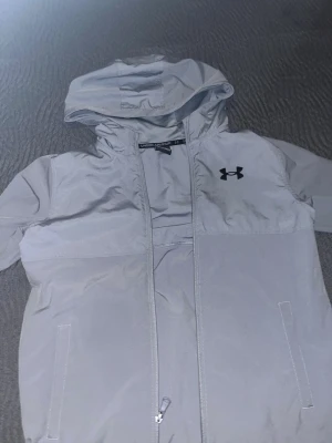 Ljusgrå vindjacka från Under Armour - Säljer en stilren ljusgrå vindjacka passar 12-13 åringar från Under Armour med huva och dragkedja framtill. Jackan har två fickor, svart logga på bröstet som har en liten bit av den som åkt av på under armour loggan på framsidan. 