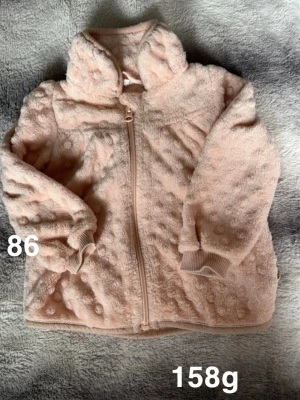 Ljusrosa fleecejacka med huva - Supermjuk ljusrosa fleecejacka med huva och dragkedja framtill. Jackan har ett bubbligt strukturmönster och ribbade muddar vid ärmsluten. Perfekt för kyliga dagar och enkel att dra på över andra kläder.🌸 Fint använt skick. Storlek 86