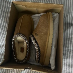 Bruna UGG slip-on boots i mocka - Säljer ett par bruna slip-on boots från UGG i mjuk mocka med fluffigt foder. Skorna har en beige, räfflad sula och dekorativt mönster runt öppningen. Perfekta för kalla dagar och riktigt sköna att ha på sig.