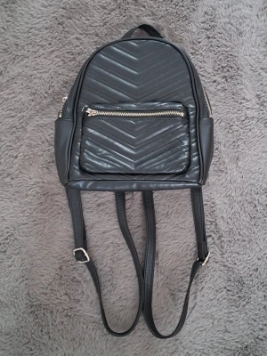Svart ryggsäck från Divided H&M - Snygg svart ryggsäck från Divided H&M i syntetmaterial med quiltad framsida och silvriga dragkedjor. Väskan har justerbara axelband, ett stort huvudfack och en praktisk ytterficka med dragkedja. Perfekt storlek för vardagens essentials.
