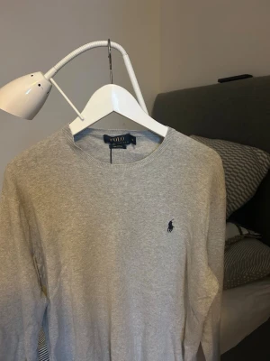 Grå långärmad tröja från Polo Ralph Lauren - Säljer en stilren grå långärmad tröja från Polo Ralph Lauren i mjuk bomull. Tröjan har rund hals och den klassiska broderade loggan i mörkblått på bröstet. Perfekt för dig som gillar enkel och clean stil.