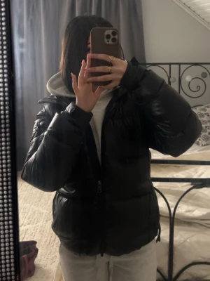 Svart pufferjacka från Berkeley - Säljer en svart pufferjacka från Berkeley med glansig finish och blått innerfoder med stort tryck. Jackan har dragkedja framtill, snörning i nederkant och långa ärmar. Perfekt för kalla dagar och riktigt skön att bära.