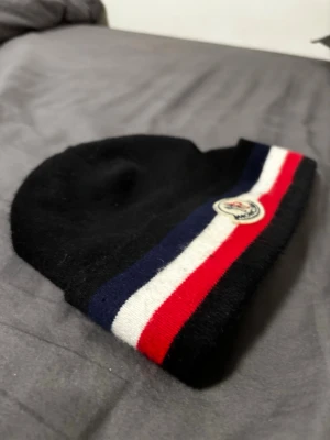 Moncler svart mössa med ränder - Snygg svart mössa från Moncler med breda ränder i marinblått, vitt och rött längs kanten. Klassisk Moncler-logga framtill. Tillverkad i mjukt material som känns lyxigt och passar perfekt till kalla dagar.