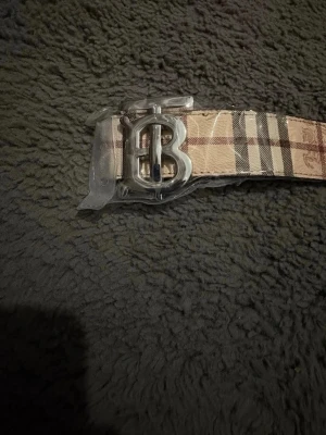 Rutig beige skärp med B-spänne - Säljer ett klassiskt Burberry-inspirerat skärp i beige med rutigt mönster och silverfärgat B-spänne. Skärpet är gjort i skinnimitation och har detaljer med ingraverade hästar. Perfekt accessoar för att lyfta din outfit.