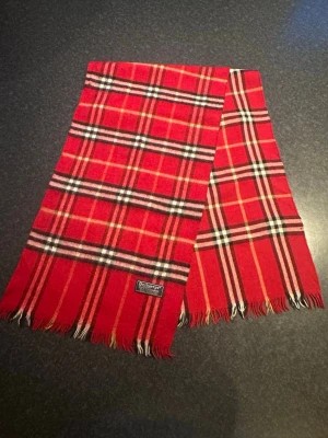 Rutig röd Burberrys halsduk i kashmir - Säljer en klassisk röd halsduk från Burberrys i 100% kashmir. Det är ett litet hål ingen men inget man märker av när man har den på.Den har ett rutigt mönster i svart, vitt och beige samt fransar i kanterna. Perfekt accessoar för att lyfta din vinterstil med lyxig känsla och tidlös design.